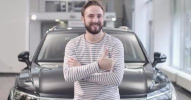 Soyunmuş süveter giymiş gülümseyen beyaz adamın portresi yeni arabanın önünde baş parmağını gösteriyor. Yeni otomobille poz veren memnun bir alıcı. Araba galerisi, araba işi. Sinema 4k görüntüleri Prores Hq.