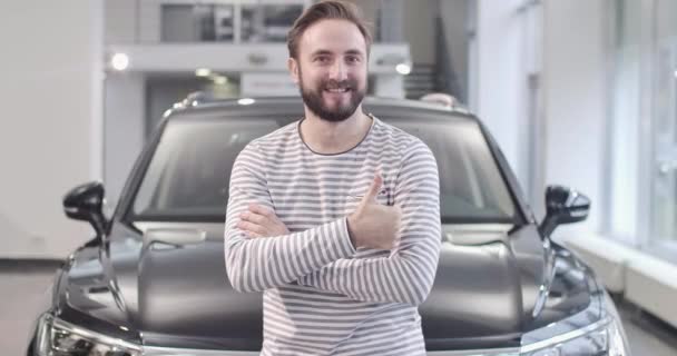 Retrato de un hombre caucásico sonriente en suéter desnudo mostrando el pulgar hacia arriba frente a un coche nuevo. Comprador satisfecho posando con un automóvil nuevo. Concesionario de autos, negocio de autos. Cinema 4k metraje ProRes HQ . — Vídeos de Stock