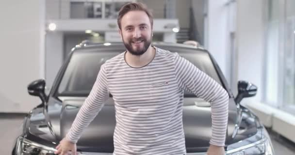 Счастливый бородатый белый парень в раздетом свитере позирует с новой черной машиной. Успешный человек покупает машину в автосалоне. Автобизнес. Съемки в кинотеатре 4k . — стоковое видео