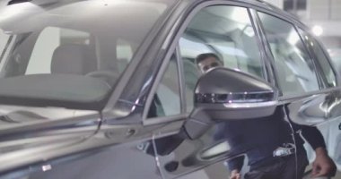 Araba galerisinde ya da otoparkta duran yeni siyah otomobilin yakın çekimi. Sakallı beyaz adam salona giriyor ve motoru çalıştırıyor. Araba işi. Sinema 4k görüntüleri Prores Hq.