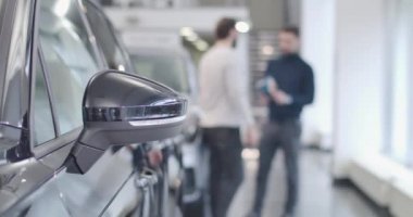 Park yerinde duran arabanın siyah kanat aynasına yakın çekim. Arka planda konuşan iki bulanık beyaz adam. Araba galerisi, araba işi. Sinema 4k görüntüleri Prores Hq.