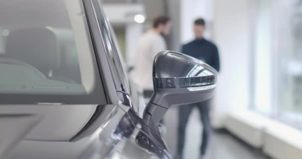 Dos hombres caucásicos borrosos con ropa casual hablando al fondo. Primer plano del espejo retrovisor lateral negro en primer plano. Concesionario de autos, negocio de autos. Cinema 4k metraje ProRes HQ . — Vídeos de Stock