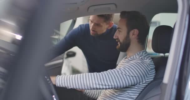 Şoför koltuğunda oturan ve erkek araba satıcısının açıklamalarını dinleyen beyaz adamın yan görüntüsü. Genç başarılı iş adamı galeride yeni bir araç alıyor. Sinema 4k görüntüleri Prores Hq — Stok video