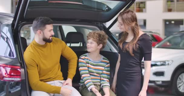 Μικρό χαριτωμένο καυκάσιο αγόρι με σγουρά μαλλιά που μιλάει με τους γονείς του στην αντιπροσωπεία αυτοκινήτων. Χαρούμενη οικογένεια που διαλέγει αυτοκίνητο μαζί. Άνθρωποι που νοικιάζουν ή αγοράζουν οχήματα. Κινηματογράφος 4k πλάνα Prores Hq. — Αρχείο Βίντεο