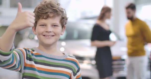 Faccia da primo piano di un bel ragazzo caucasico con i capelli ricci che mostrano il pollice in alto. I suoi giovani genitori parlano in background nella concessionaria di auto. La famiglia compra un'automobile nuova. Cinema 4k riprese ProRes HQ . — Video Stock