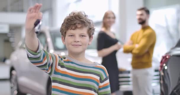 Cute Caucasian boy with curly hair showing car keys and thumb up. Blurred young parents talking at the background. Successful family buying new vehicle. Cinema 4k footage ProRes HQ. — ストック動画