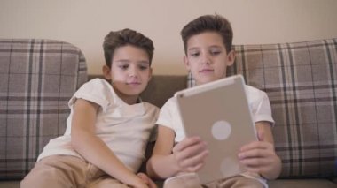 Tablet ekrana bakıp gülümseyen iki beyaz ikiz kardeşin ön görüntüsü. Kardeşler evdeki rahat kanepede dinleniyor..