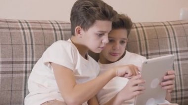 Tablette video oyunu oynayan ve beşlik çakan iki beyaz ikizin yakın plan portresi. Kardeşler içeride birlikte vakit geçiriyorlar. Kardeşler evde dinleniyor..