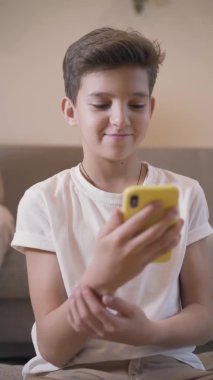 Akıllı telefon kullanan Kafkas esmer çocuğun portresi. Evde sosyal medyayı kullanan beyaz tişörtlü bir öğrenci. İnternet bağımlılığı, sosyal medya bağımlılığı. Dikey çekim.