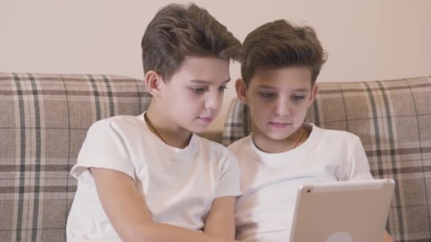 Facce da primo piano di fratelli gemelli caucasici che giocano ai videogiochi su tablet e danno il cinque. I fratelli si divertono insieme in casa. Gemelli a riposo a casa . — Video Stock