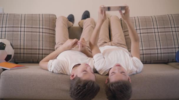Two cheerful Caucasian twin brothers laying on couch and using tablet. Siblings resting together at home. Children using social networks, game addiction, modern kids. — ストック動画
