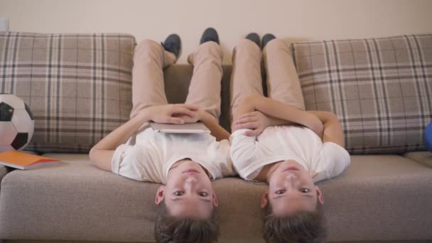 Two Caucasian twin brothers laying laying upside down on sofa, looking at camera and laughing. Cheerful siblings resting together at home. Modern kids having fun indoors. — ストック動画