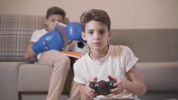 Close-up portrait of cute Caucasian boy playing game console as his twin brother sitting at the background in boxing gloves and using tablet. Cheerful siblings enjoying free time together at home. — Stock Video
