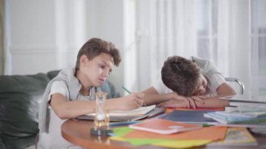 Masada oturan ve yazı yazan Kafkasyalı okul çocuğunun yan görüntüsü. Yorgun ikiz kardeşi egzersiz kitabının üzerine yatmış ve uykuya dalmış. Çocuk kardeşini uyandırdı ve onu çalışmaya zorladı..