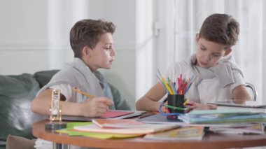 Masada oturup ders çalışan iki beyaz ikiz kardeş. Bir çocuk hile yapmaya çalışıyor, kardeşi egzersiz defterini kapatıyor. Okul çocukları evde ödev yapıyor. Eğitim kavramı.