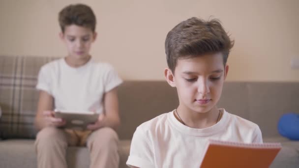 Retrato de close-up do jovem estudante lendo livro, voltando-se para trás e olhando para seu irmão gêmeo sentado com tablet no fundo. Os irmãos estudam juntos em casa. Conceito de educação, aprendizagem . — Vídeo de Stock