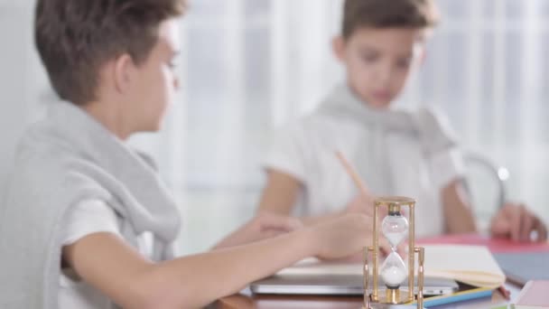 Portret van twee wazige Kaukasische tweelingbroers die op de achtergrond high five gaven als gedraaid zandglas dat op de voorgrond aan tafel verbleef. Succesvolle schooljongens die samen huiswerk maken thuis. — Stockvideo