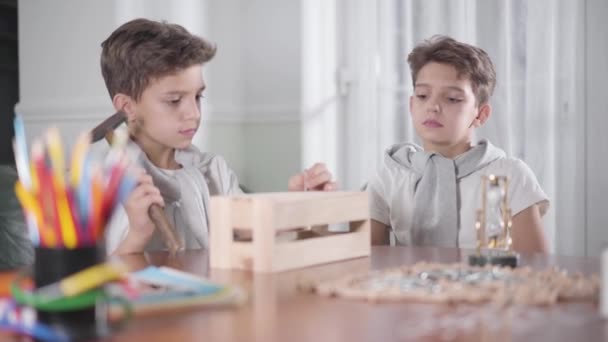 Retrato de dos hermanos gemelos caucásicos haciendo carpintería. Un chico girando vidrio de arena y su hermano empezando a martillar clavos. Carpintería para niños, hobbies . — Vídeos de Stock