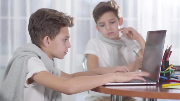 Portret van een blanke schooljongen die naar zijn tweelingbroer kijkt die spelletjes speelt op laptop en zucht. Brunette jongen gebaart ja en typt weer. Verschillende levensstijlen, internetverslaving, sociale media. — Stockvideo