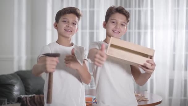 Radostní bělošští bratři s dvojčaty, ukazující před kamerou kladivo a dřevo a usmívající se. Sourozenci se chlubí svou prací. Zaměření se změní z pozadí do popředí. Dřevoobrábění, tesařství pro děti, hobby. — Stock video