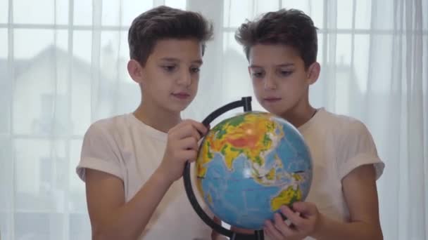 Close-up portret van brunette Kaukasische schooljongen met wereldbol en uitleg over aardrijkskunde aan zijn tweelingbroer. Broers en zussen doen samen huiswerk. Onderwijs concept, intelligentie. — Stockvideo