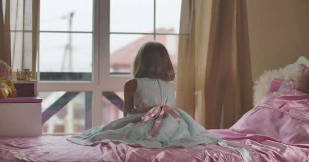 Vue arrière de la petite fille caucasienne assise sur le lit et regardant par la fenêtre. Mignon enfant se tournant vers la caméra et tenant son visage avec la main. Charmante fille reposant dans sa chambre à la maison . — Video
