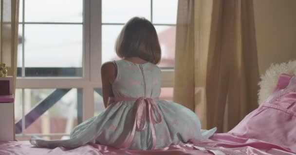 Close-up van het blanke meisje dat op het bed zit en over de schouder naar de camera kijkt. Kind in jurk met vlinders zittend op roze bed in haar kamer thuis. — Stockvideo