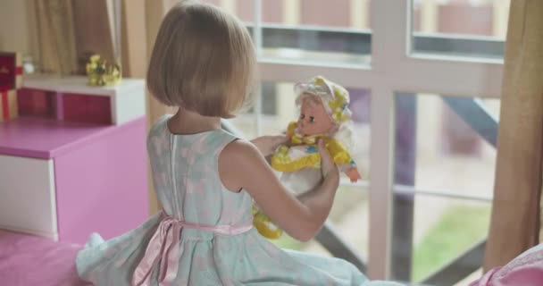 Bovenaanzicht van het kleine blanke blonde meisje dat met pop speelt als zittend aan een roze bed. Kind in mooie blauwe jurk met roze vlinders knuffelen het speelgoed en de vaststelling van zijn hoed. Jeugd, vrije tijd. — Stockvideo