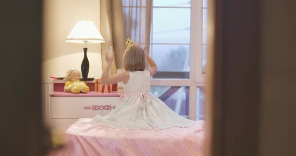 Schießen durch Tür von kaukasischen Mädchen in schönem Kleid sitzt auf rosa Bett und setzt Spielzeug Krone auf. Kleine Dame zieht sich an und schaut in den Spiegel. — Stockvideo