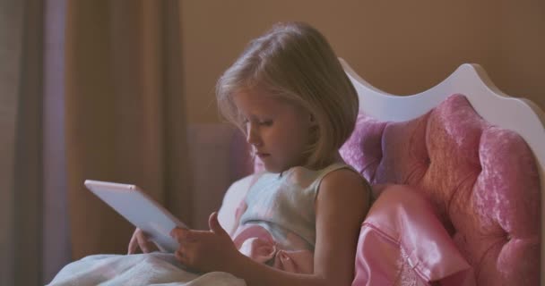 Blond Caucasian girl lying aside tablet and going to bed. Pretty child falling asleep in pink bed. Resting at home, carefree childhood. — Stock Video