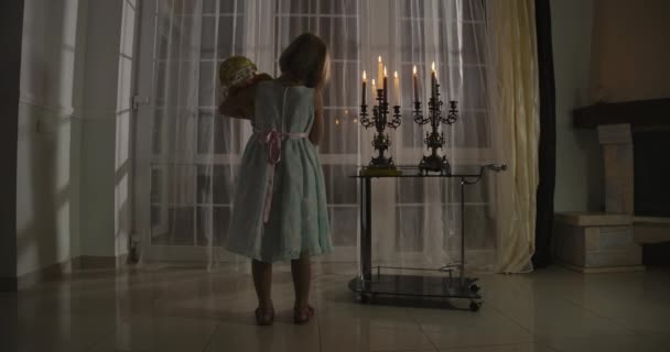 Vue arrière de la petite fille caucasienne debout à la chandelle avec poupée. Enfant caressant son jouet et regardant la fenêtre. Peur, peur, étrangeté . — Video