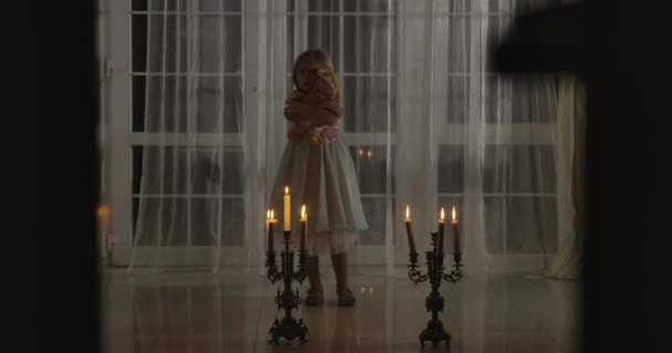 Konstig vit tjej som står i levande ljus med docka. Skrämmande barn som ser sig omkring. Skrämsel, rädsla, konstighet. — Stockvideo