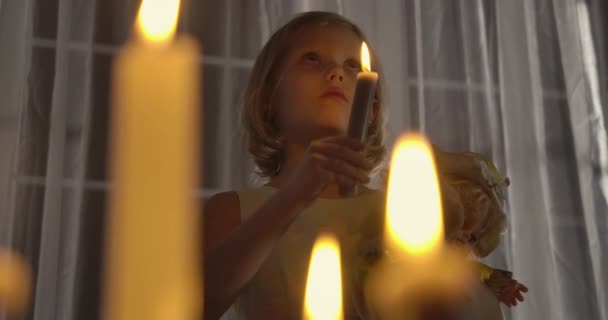 Close-up rosto de menina branca estranha segurando boneca e vela e olhando para cima. Criança estranha em pé à luz de velas na frente de uma grande janela à noite. Medo, medo, estranheza . — Vídeo de Stock