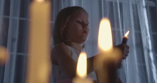 Retrato de menina caucasiana segurando boneca e soprando vela. Criança estranha em pé à luz de velas na frente de uma grande janela à noite. Medo, medo, estranheza . — Vídeo de Stock