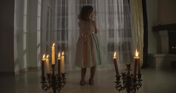 Macchina fotografica in avvicinamento piccola ragazza caucasica in piedi a lume di candela e tenendo bambola. Strana bambina che accarezza il suo giocattolo e si guarda intorno. Paura, paura, stranezza . — Video Stock