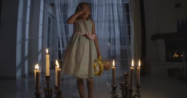 Portret van een bang blank meisje in een mooie jurk met een pop, die haar ogen sluit met de hand. Bang kind bij kaarslicht, wind waait door witte gordijnen op de achtergrond. — Stockvideo
