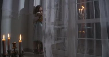 Little Caucasian girl standing in the corner in front of big window and holding doll. Child in candlelight playing with her toy. Scare, fear, strangeness.