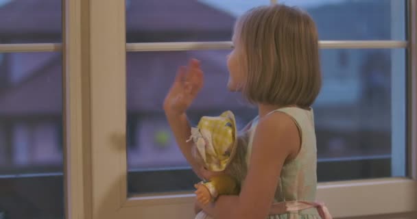 Feliz niña caucásica sosteniendo la muñeca y saludando por la ventana. Niño despedir a los huéspedes o familiares por la noche. Elegante niño en vestido hermoso pasar tiempo en casa . — Vídeos de Stock