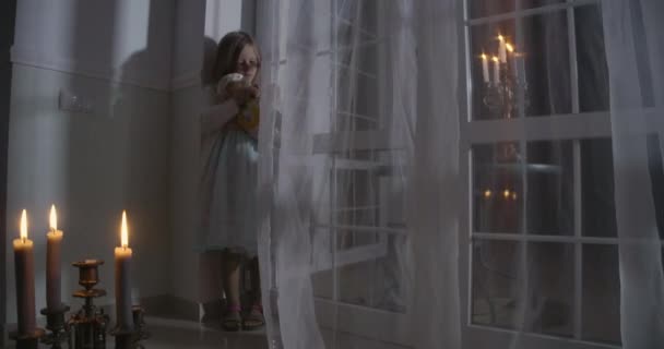 Little Caucasian girl standing in the corner in front of big window and holding doll. Child in candlelight playing with her toy. Scare, fear, strangeness. — 图库视频影像