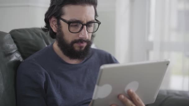 Portret van een geconcentreerde blanke man in een bril met een tablet. Jonge bebaarde zakenman die in het weekend thuis met problemen omgaat. Afstandelijk werk, freelance, overwerk. — Stockvideo