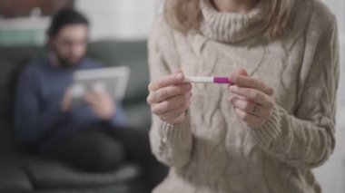İki eliyle hamilelik testi yapıp göğsüne koyan beyaz bir kadının yakın çekimi. Arka planda koltukta oturan bulanık sakallı adam tablet kullanıyor. Mutlu kız, hamilelik.