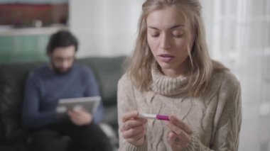 Kahverengi gözlü, düşünceli, sarışın, beyaz bir kız gebelik testi yapıyor ve arka planda tablet kullanan bir adama bakıyor. Erkek arkadaşına ya da kocasına hamileliğini nasıl anlatacağına karar veren şok olmuş bir kadın..