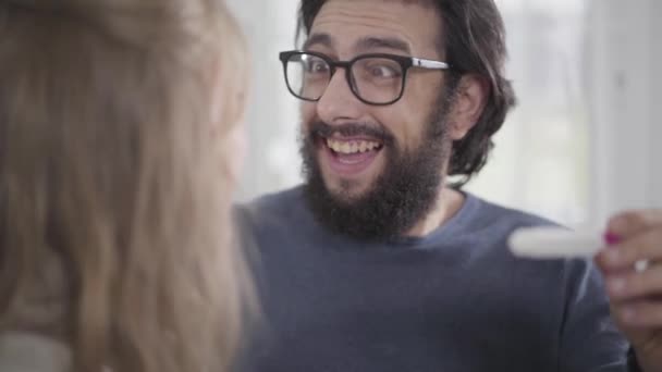 Gros plan sur le visage du jeune homme caucasien avec la barbe noire imitant la joie comme sa femme montrant test de grossesse affirmative, et câlin fille. Choqué mec avec l'expression de la peur sur le visage . — Video