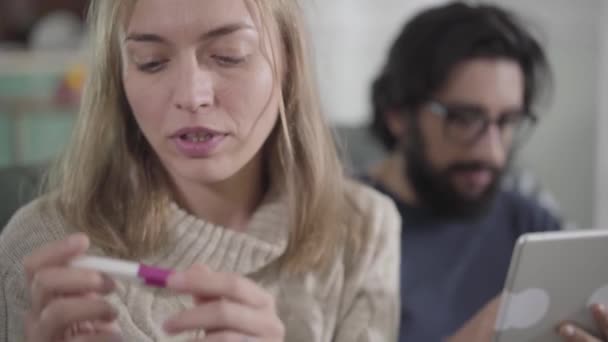 Close-up face of sad blond Caucasian girl with brown eyes holding pregnancy test and looking back at man using tablet at the background. Young woman deciding how to tell husband about pregnancy. — Stock Video