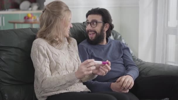 Närbild av kaukasiska blond kvinna visar sin pojkvän eller make bekräftande graviditetstest. Skäggig man kramar flickan och ler. Lycklig ung familj väntar på barn. Graviditet, förväntan. — Stockvideo