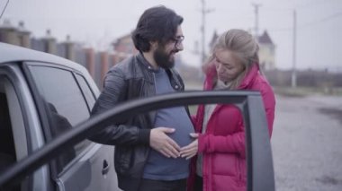 Gülümseyen beyaz sarışın kadın kocasını okşuyor hamile karnını okşuyor ve bebek vuruşlarını taklit ediyor. Sosyal rollerini paylaşan neşeli bir çift. Şaka, komik..