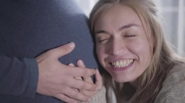 Çekici, beyaz bir kadının yakın plan yüzü hamile erkeklerin karnına kulak veriyor. Bebeğin kalp atışlarını dinleyen pozitif bir kız. Sosyal rollerini değiştiren komik bir çift. Şaka, eğlence, şaka.