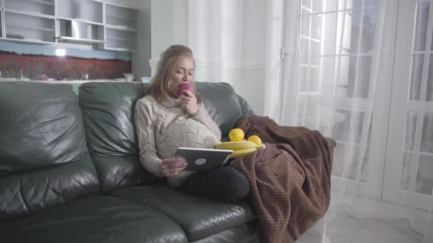 Portret van een zwanger vrolijk meisje dat video kijkt op tablet en appel eet. Jonge blanke vrouw die thuis op de bank rust. Zwangerschap, zwangerschap, zwangerschapsvreugde, gezond eten. — Stockvideo