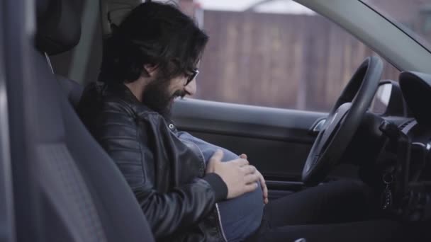 Pohled zblízka na legračního těhotného muže sedícího na sedadle řidiče a hladícího si břicho. Portrét usměvavého dospělého chlapa s kudrnatým černým vousem a brýlemi. Vtipkování, zábava. — Stock video