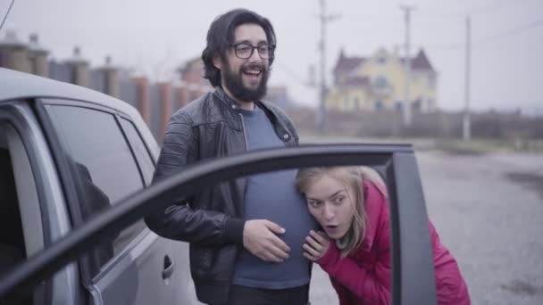 Positive bearded Caucasian man in eyeglasses imitating baby hitting as her girlfriend or wife holding ear next to his pregnant belly. Funny family exchanging their social roles. Joking, fun. — Stock Video